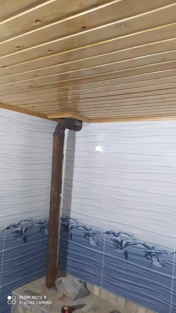 Drewniana boazeria sufitowa z rurą przechodzącą przez nią w rogu pomieszczenia, białe płytki z niebieskim wzorem na ścianach.