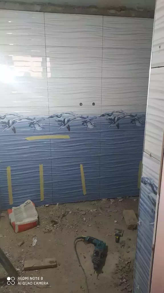 Wnętrze łazienki w trakcie układania płytek ściennych: białe płytki na górze, niebieskie z motywem delfinów na dole, widoczny worek z zaprawą i wiertarka na podłodze.