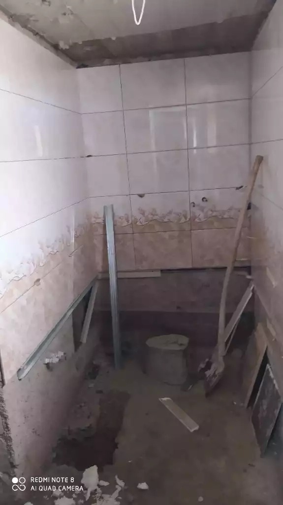 Wnętrze łazienki w trakcie remontu: częściowo położone białe płytki na ścianach, widoczne metalowe profile, wiadro z zaprawą i łopata.