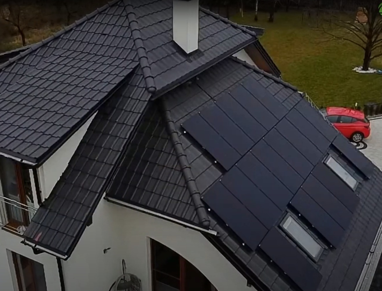 Czarny dach z dachówki ceramicznej z panelami słonecznymi zintegrowanymi z połacią, widok z góry na dom z białymi ścianami i czerwonym samochodem na podjeździe.