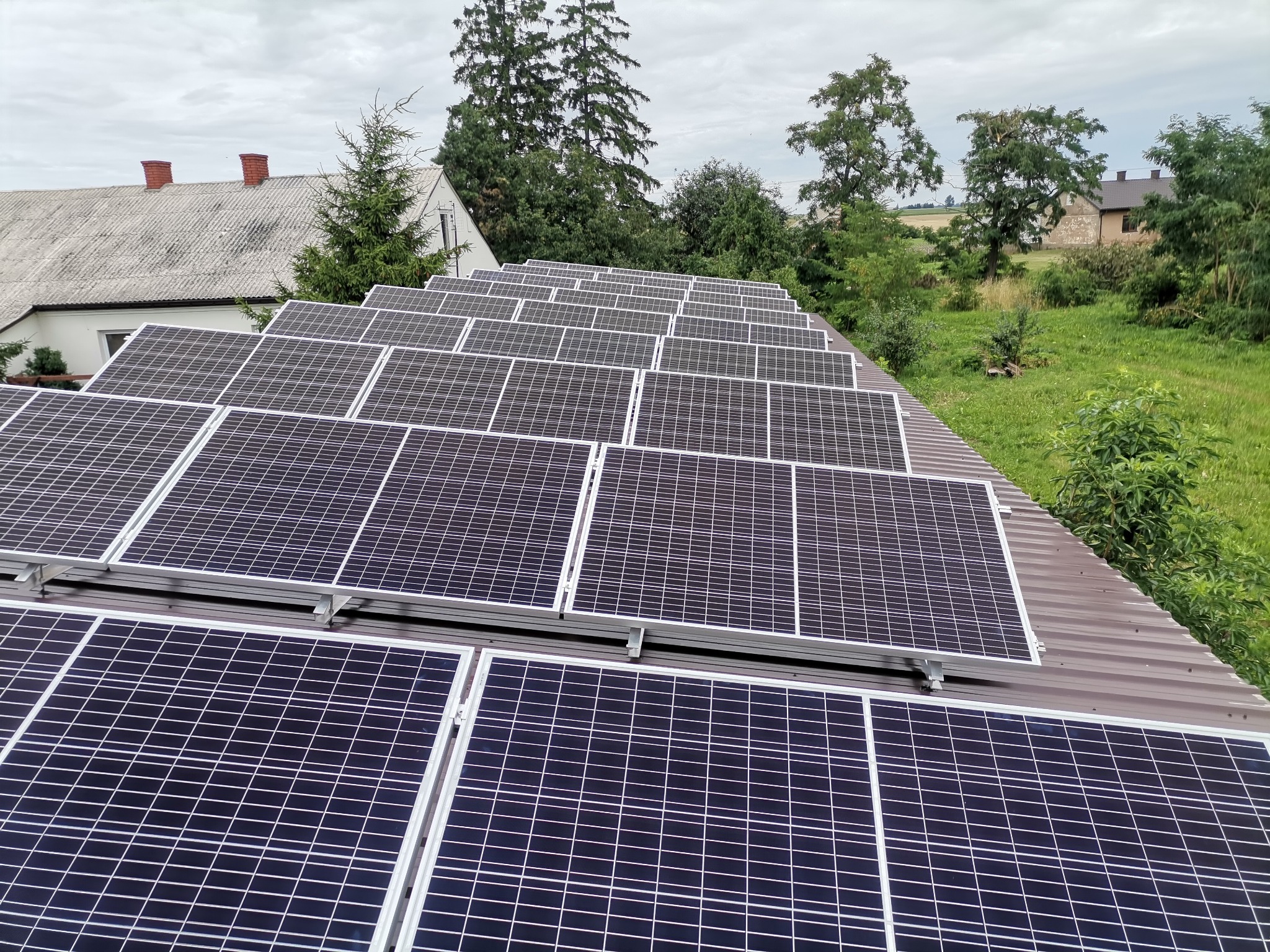 Rząd paneli słonecznych na brązowym dachu budynku gospodarczego, w tle zieleń drzew i łąk pod pochmurnym niebem.