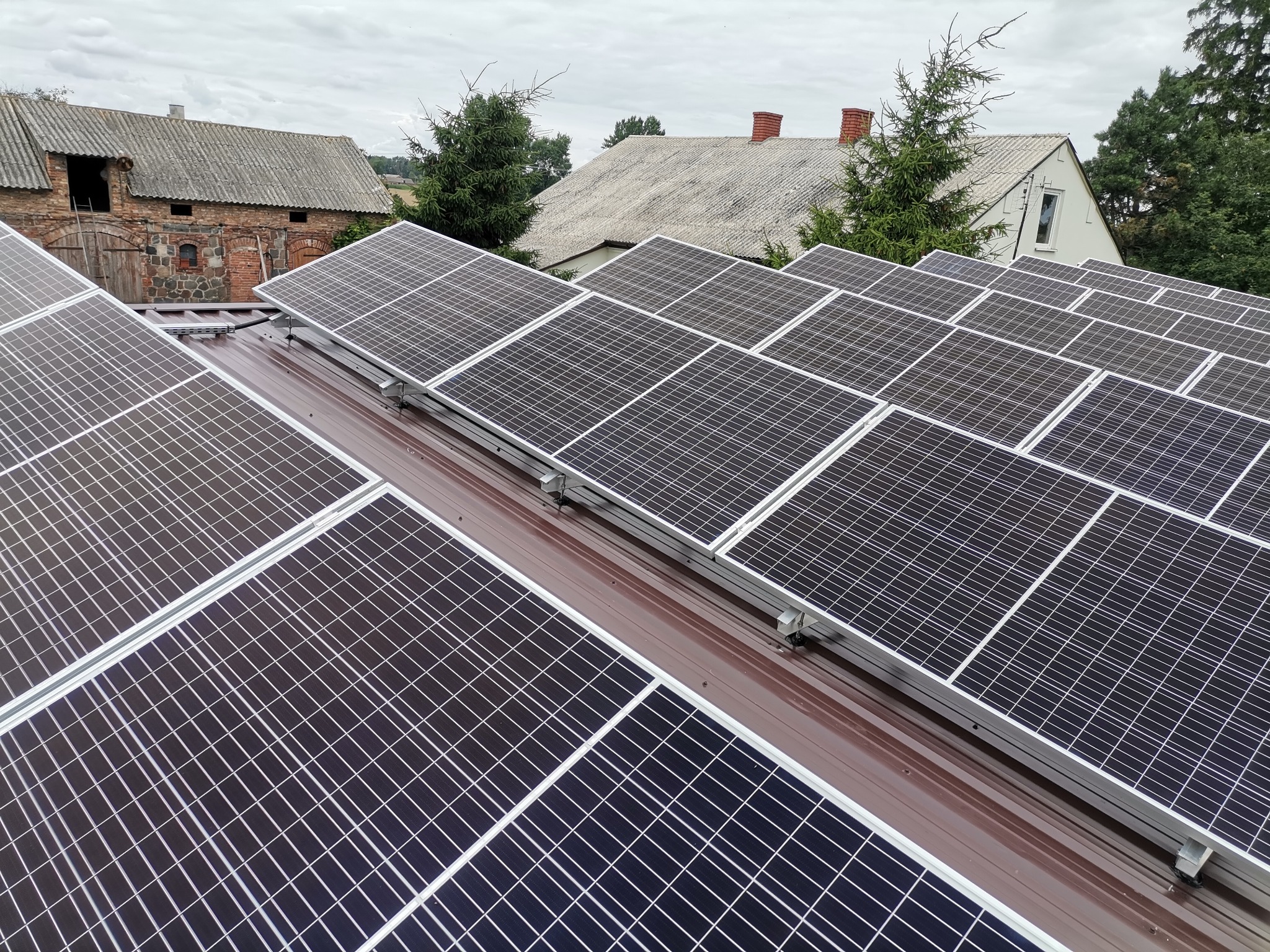 Rząd paneli słonecznych na brązowym dachu z widokiem na budynki gospodarcze i domy mieszkalne w tle, pochmurne niebo.