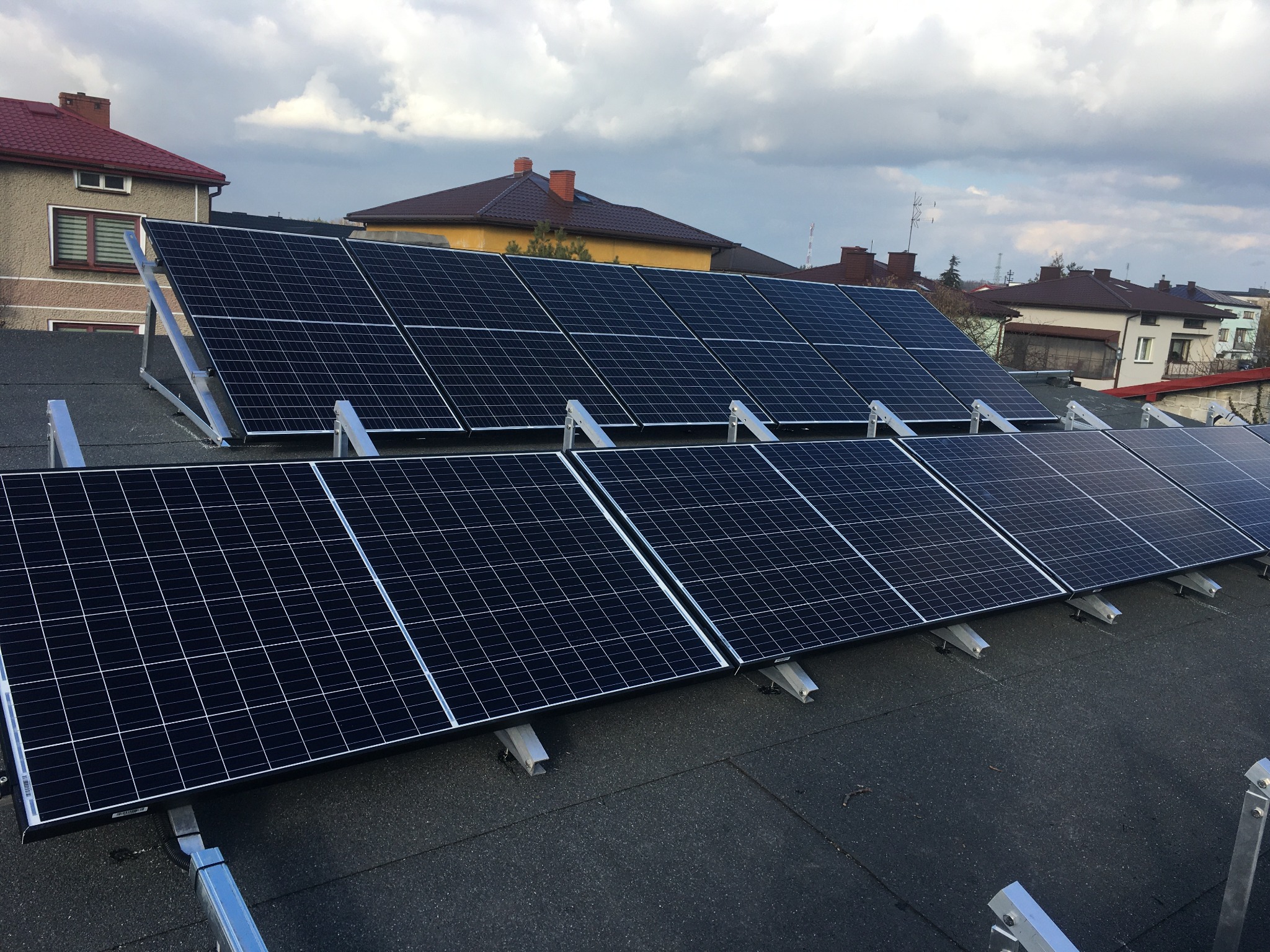 Panele fotowoltaiczne zamontowane na płaskim dachu budynku mieszkalnego, widoczne metalowe konstrukcje wsporcze i fragmenty kabli.