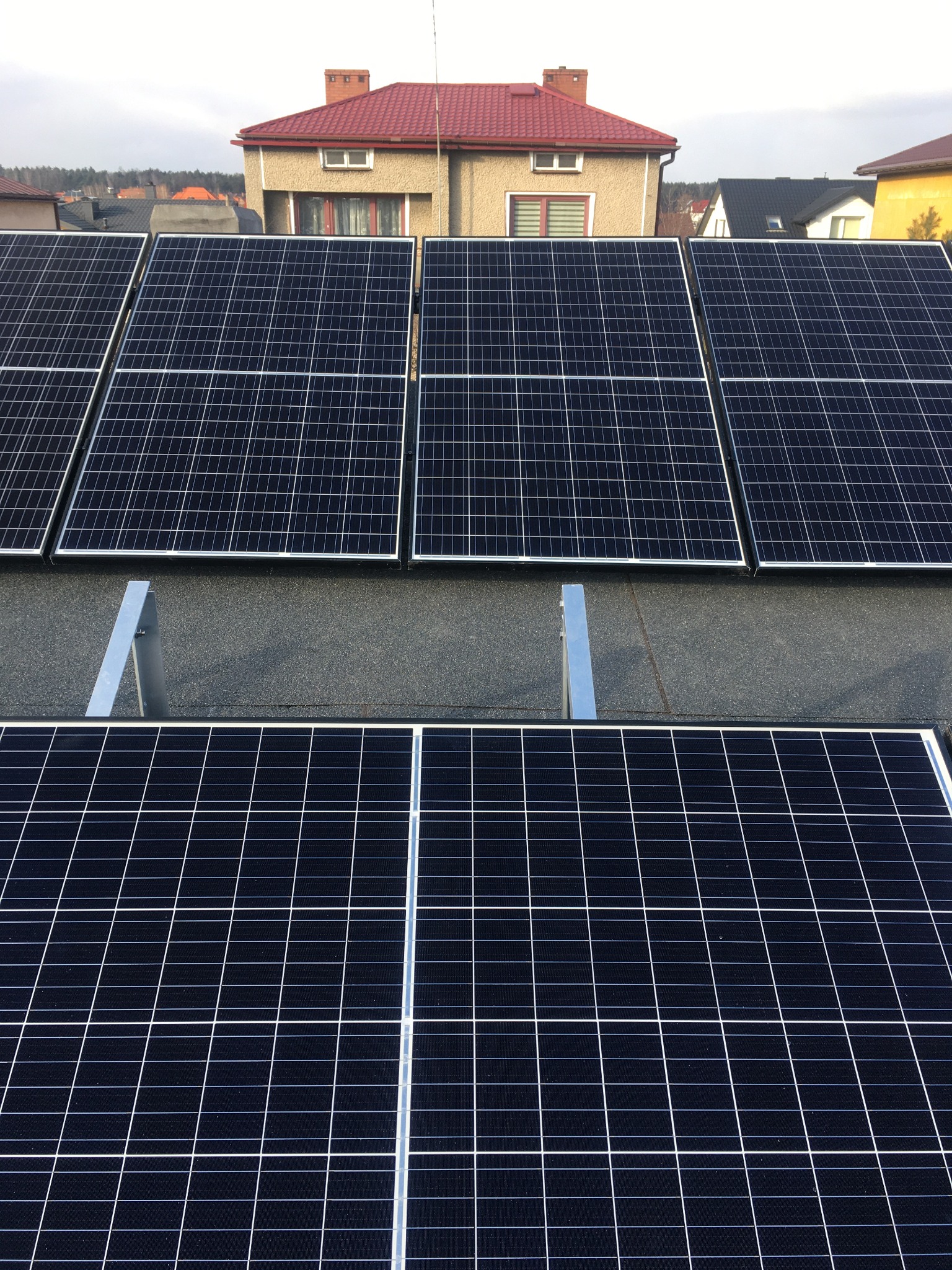 Panele słoneczne na dachu domu jednorodzinnego z czerwonym dachem, widoczne szare metalowe wsporniki montażowe.