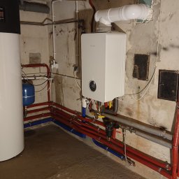 EUROTECH Spółka z o.o. - Biały, kondensacyjny kocioł gazowy marki Bosch zamontowany na ścianie z widocznymi rurami instalacji grzewczej, obok stoi biały zasobnik ciepłej wody marki Buderus, widoczne liczne rury...