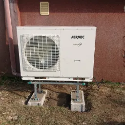 Biała jednostka zewnętrzna pompy ciepła marki Aermec, zamontowana na metalowej konstrukcji wspartej na betonowych bloczkach, na tle ściany budynku z brązowym tynkiem strukturalnym i uszkodzonym...