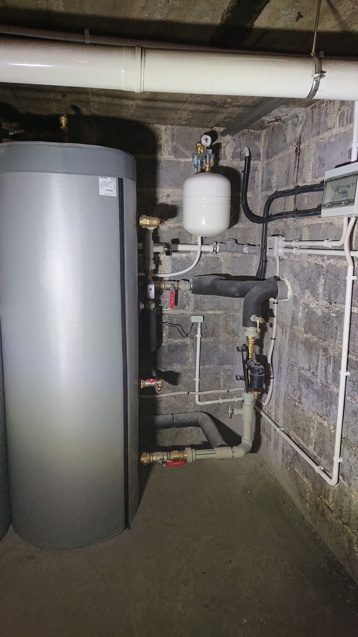 Szary zbiornik akumulacyjny z podłączonymi rurami i zaworami w piwnicy z betonowych pustaków, widoczny biały zbiornik przeponowy z manometrem i instalacja elektryczna.