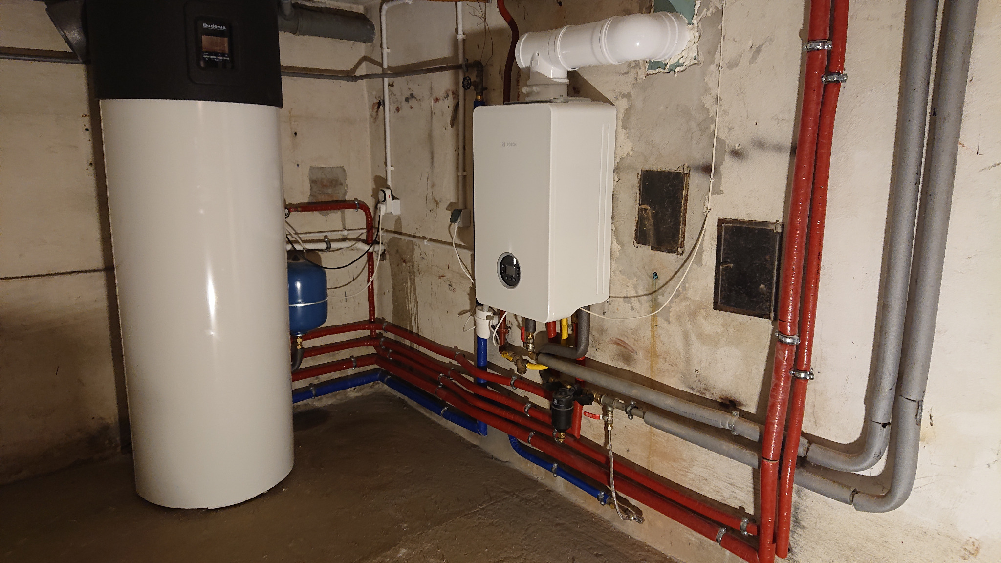 Biały, kondensacyjny kocioł gazowy marki Bosch zamontowany na ścianie z widocznymi rurami instalacji grzewczej, obok stoi biały zasobnik ciepłej wody marki Buderus, widoczne liczne rury...