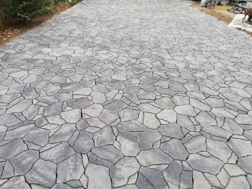 Ułożona nawierzchnia z szarej kostki o nieregularnym kształcie, widok z perspektywy, z zapasem kostki widocznym w tle po prawej stronie.