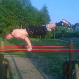 Mężczyzna bez koszulki wykonuje ćwiczenie równowagi na czerwonych poręczach w parku; w tle drzewa i domy.