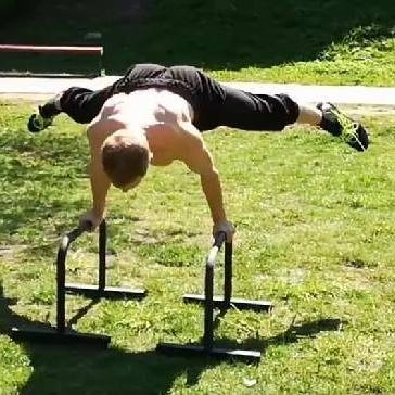 Mężczyzna bez koszulki wykonuje ćwiczenie równowagi na poręczach na trawie, nogi rozwarte w szpagacie, widok z góry.