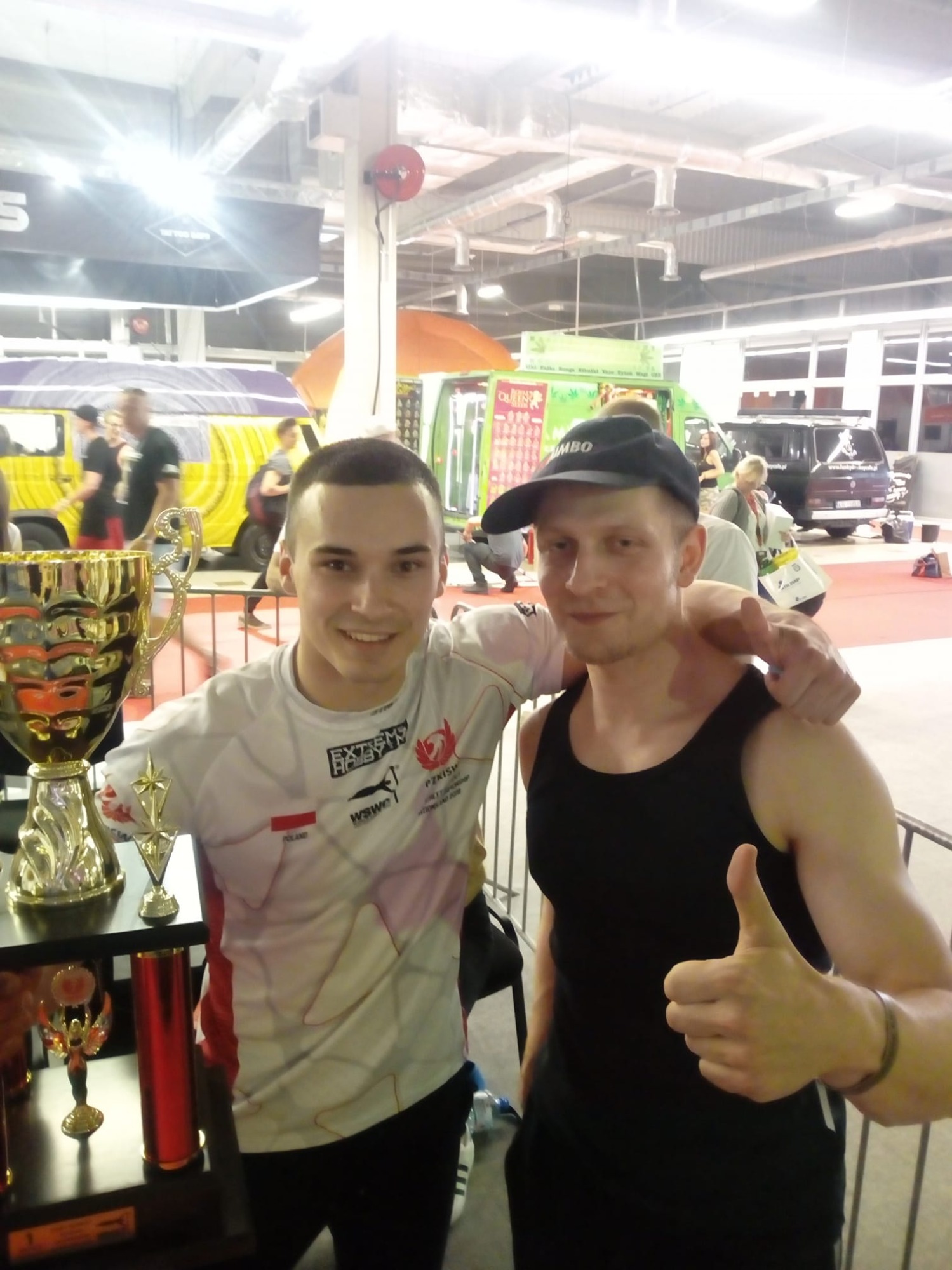 Dwóch mężczyzn pozuje z trofeami w tle hali sportowej, jeden z nich pokazuje kciuk w górę.