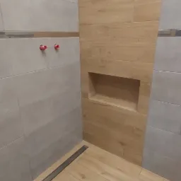 Wykończona łazienka z drewnopodobnymi płytkami, wnęką w ścianie wyłożonej tym samym materiałem oraz odpływem liniowym w podłodze.