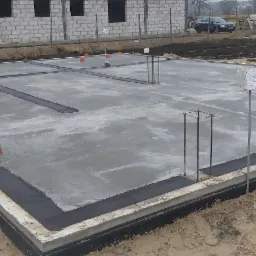 Wylany fundament z izolacją poziomą i pionową, widoczne wystające pręty zbrojeniowe oraz rury instalacyjne, w tle budynek w stanie surowym otwartym.