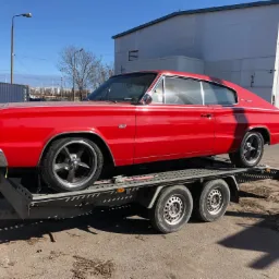 Czerwony, klasyczny samochód Dodge Charger z 1966 roku na niskopodwoziowej przyczepie lawetowej. Widoczny budynek przemysłowy w tle.