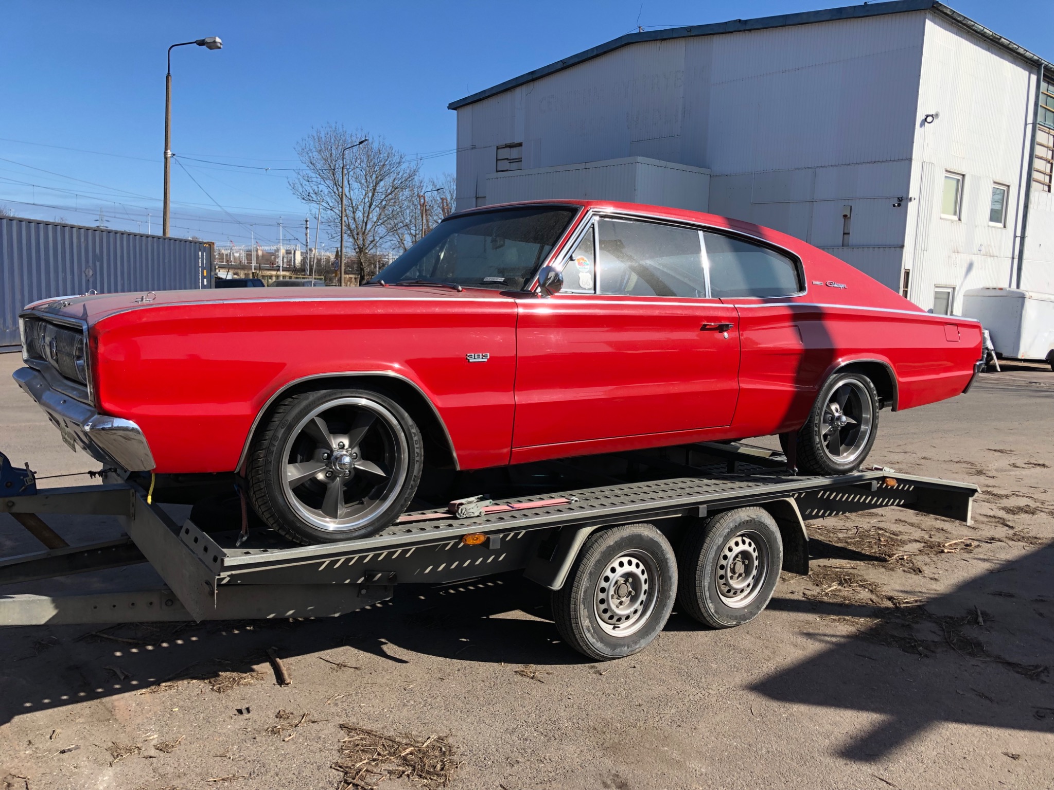Czerwony, klasyczny samochód Dodge Charger z 1966 roku na niskopodwoziowej przyczepie lawetowej. Widoczny budynek przemysłowy w tle.
