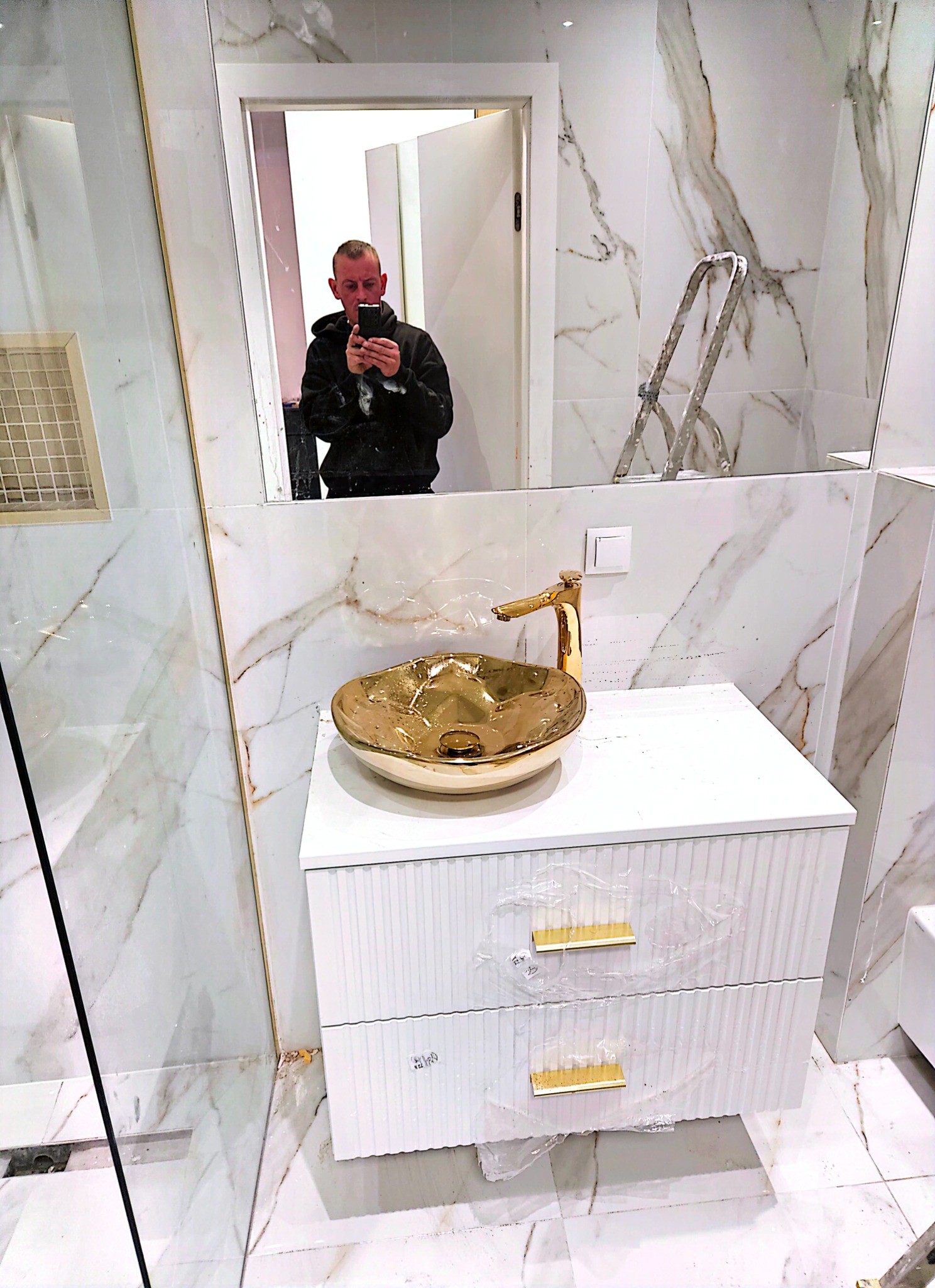 Nowoczesna łazienka w trakcie wykańczania: złota umywalka na białej szafce z ryflowanymi frontami, marmurowe płytki na ścianach, widoczne ślady po remoncie i odblask osoby robiącej zdjęcie w lustrze.