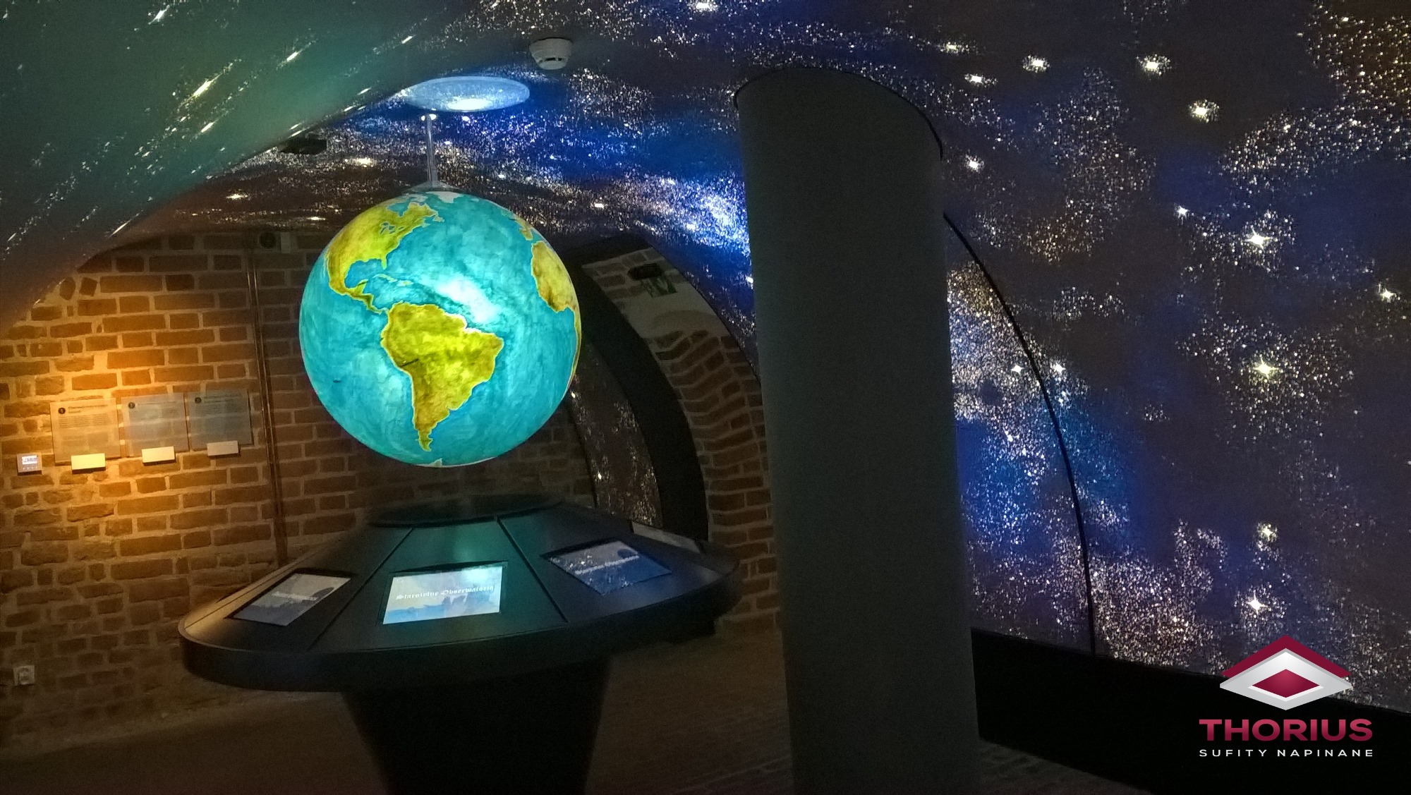Instalacja sufitu napinanego imitującego rozgwieżdżone niebo w pomieszczeniu z ceglanymi ścianami i podświetlonym modelem Ziemi.