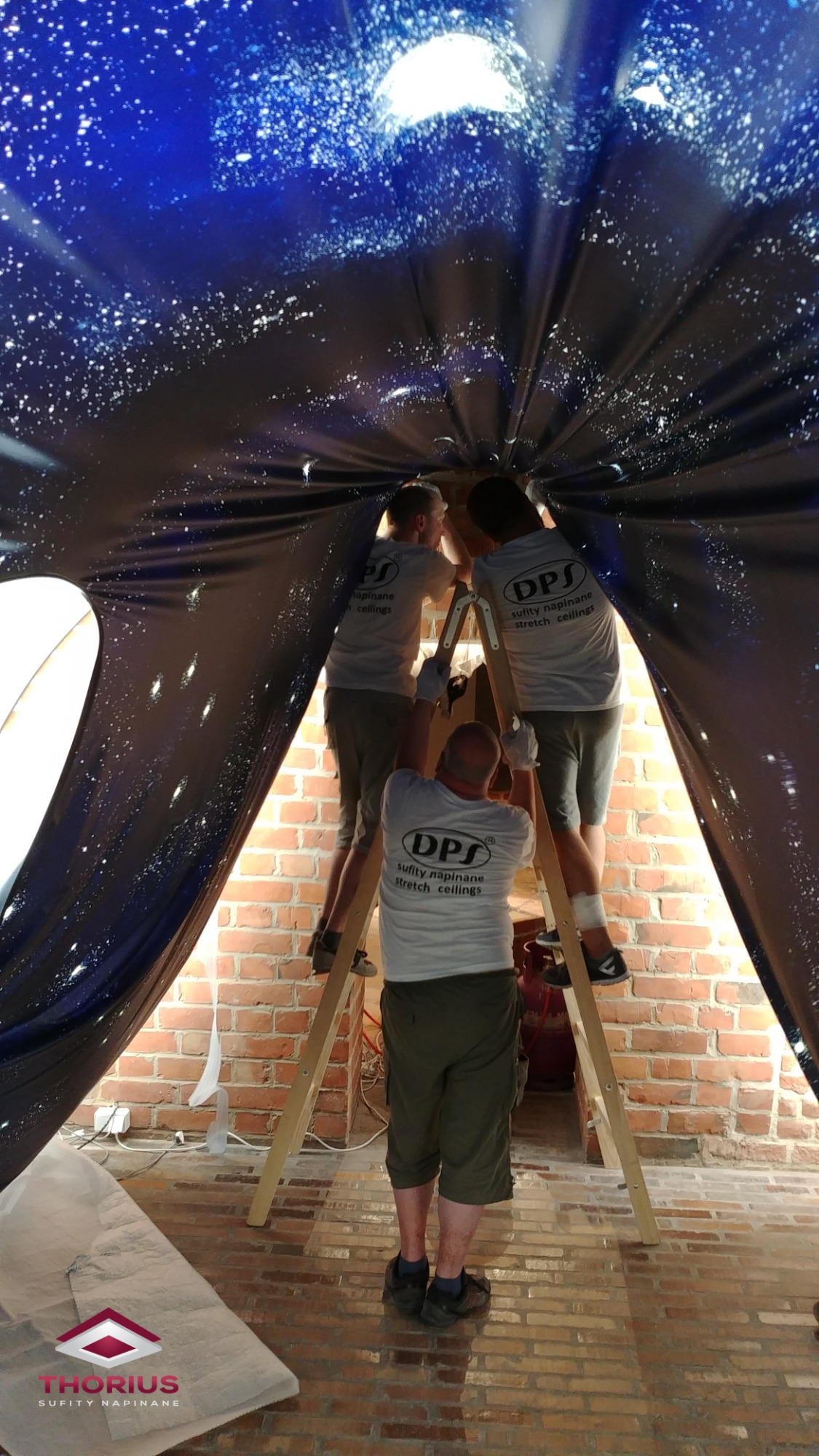 Trzech mężczyzn na drabinie montuje napinany sufit z motywem kosmosu w pomieszczeniu z ceglanymi ścianami; widoczne logo firmy na koszulkach pracowników i logo firmy na podłodze.
