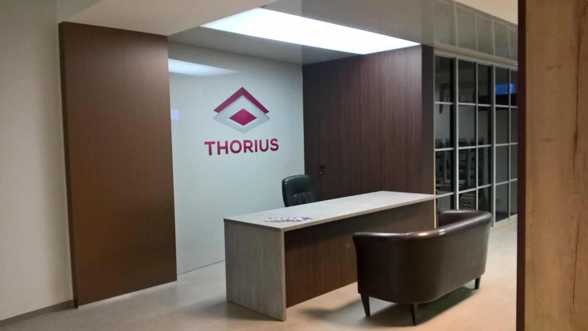 Recepcja biura z logo firmy 'Thorius' na ścianie, biurkiem z blatem w kolorze jasnego drewna, ciemnoszarym fotelem i przeszkleniem w tle.