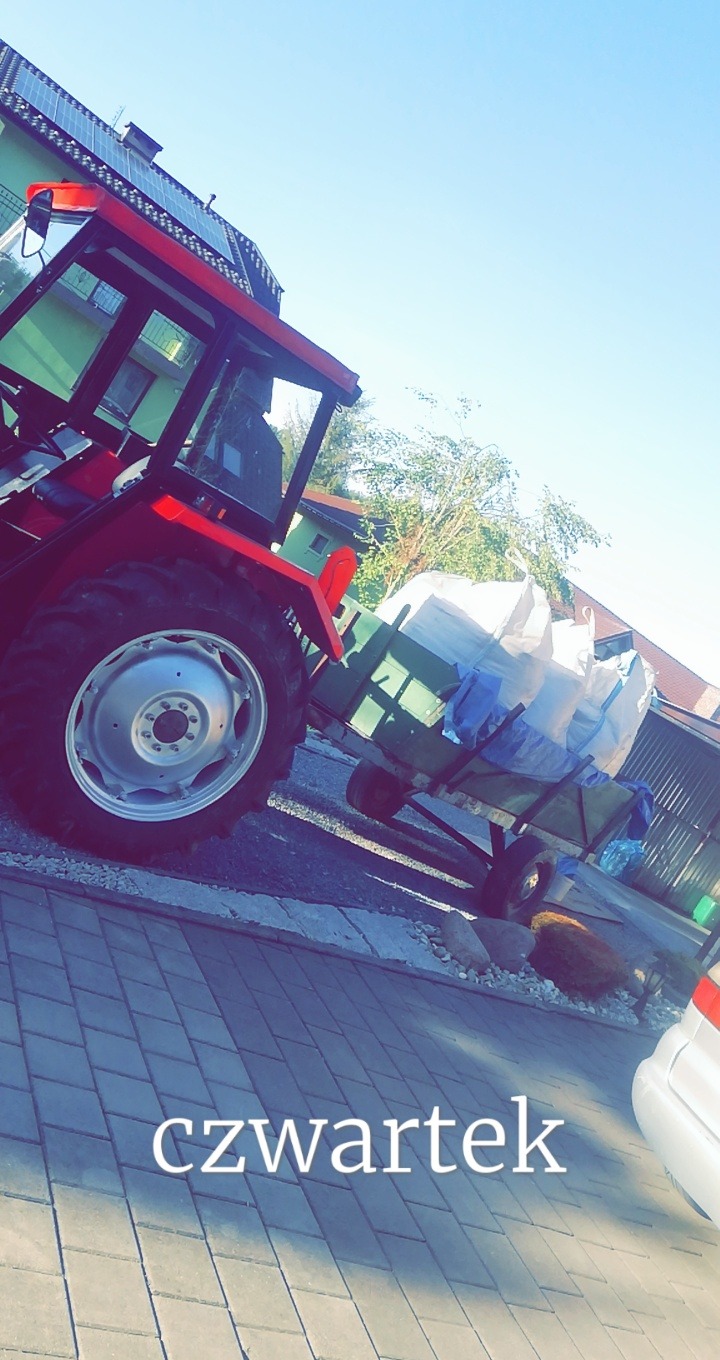 Czerwony traktor z przyczepą wypełnioną workami, prawdopodobnie na podwórku z kostki brukowej.