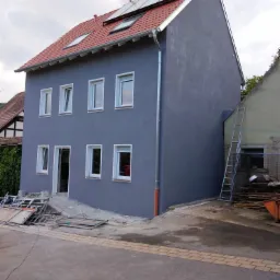 Dwupiętrowy dom z szarą elewacją po tynkowaniu, czerwonym dachem z panelami słonecznymi i białymi oknami, widoczne rusztowania i materiały budowlane przed budynkiem.