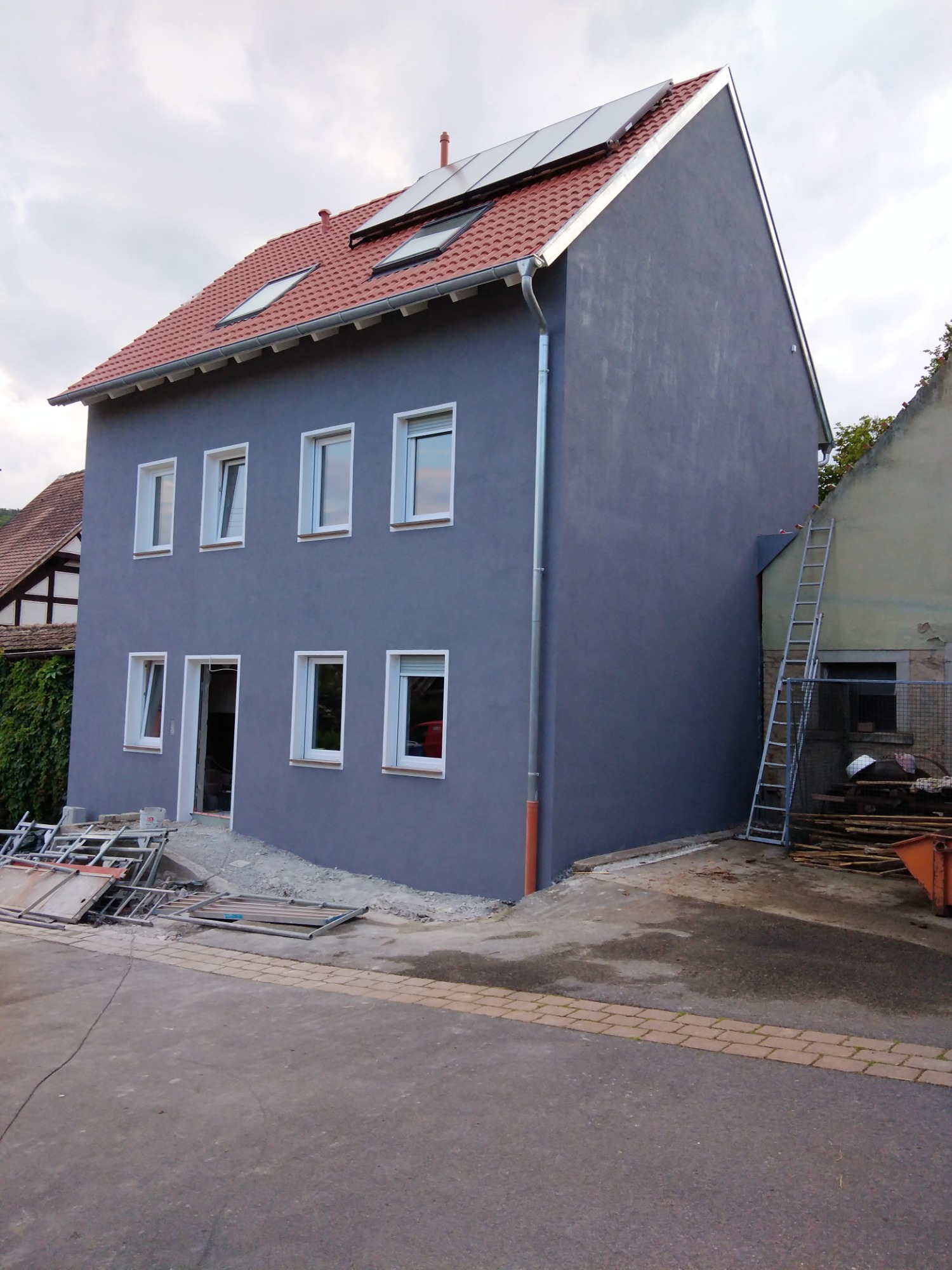 Dwupiętrowy dom z szarą elewacją po tynkowaniu, czerwonym dachem z panelami słonecznymi i białymi oknami, widoczne rusztowania i materiały budowlane przed budynkiem.