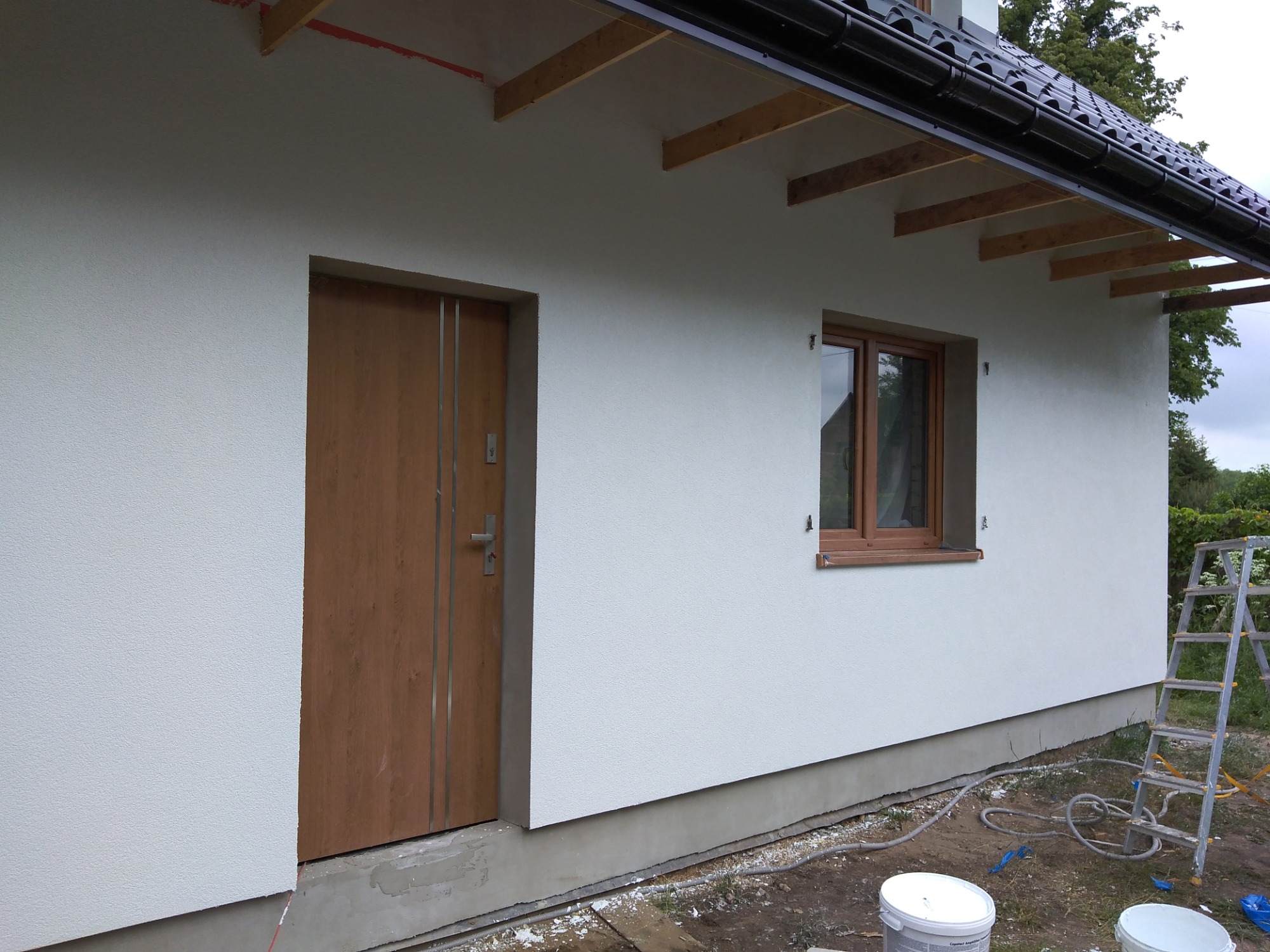 Świeżo otynkowana elewacja domu z drewnianymi elementami konstrukcyjnymi pod dachem, brązowymi drzwiami wejściowymi i oknem, widoczna drabina i wiaderka z farbą w tle.