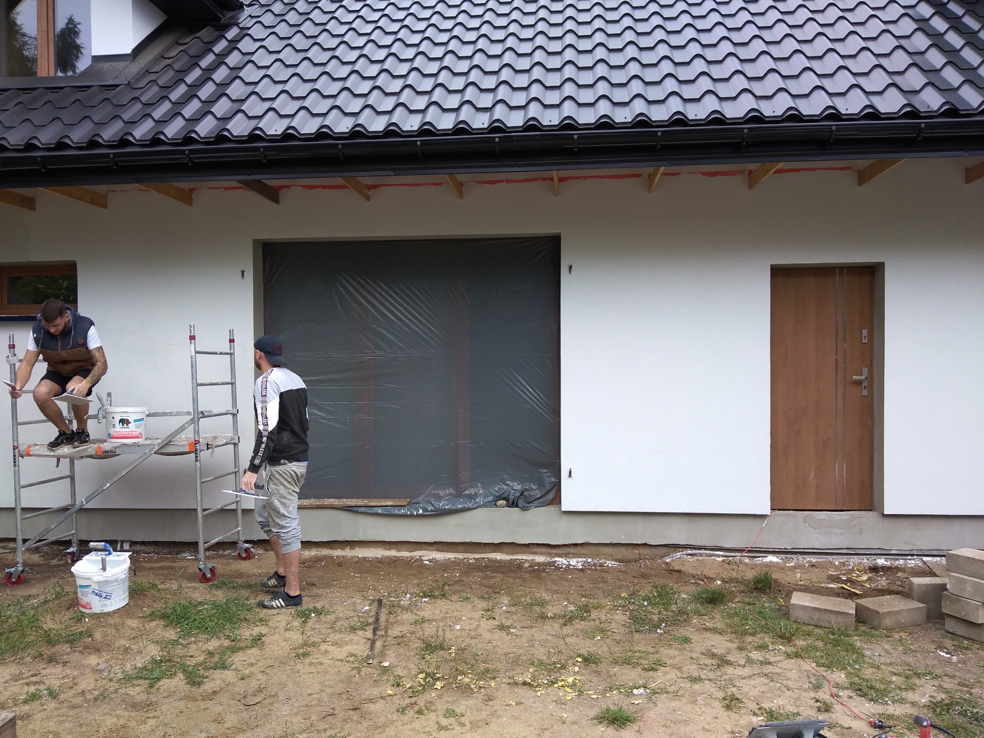 Dwóch pracowników na zewnątrz domu podczas nakładania tynku na elewację; jeden stoi na rusztowaniu, drugi na ziemi z narzędziami, okno zabezpieczone folią.