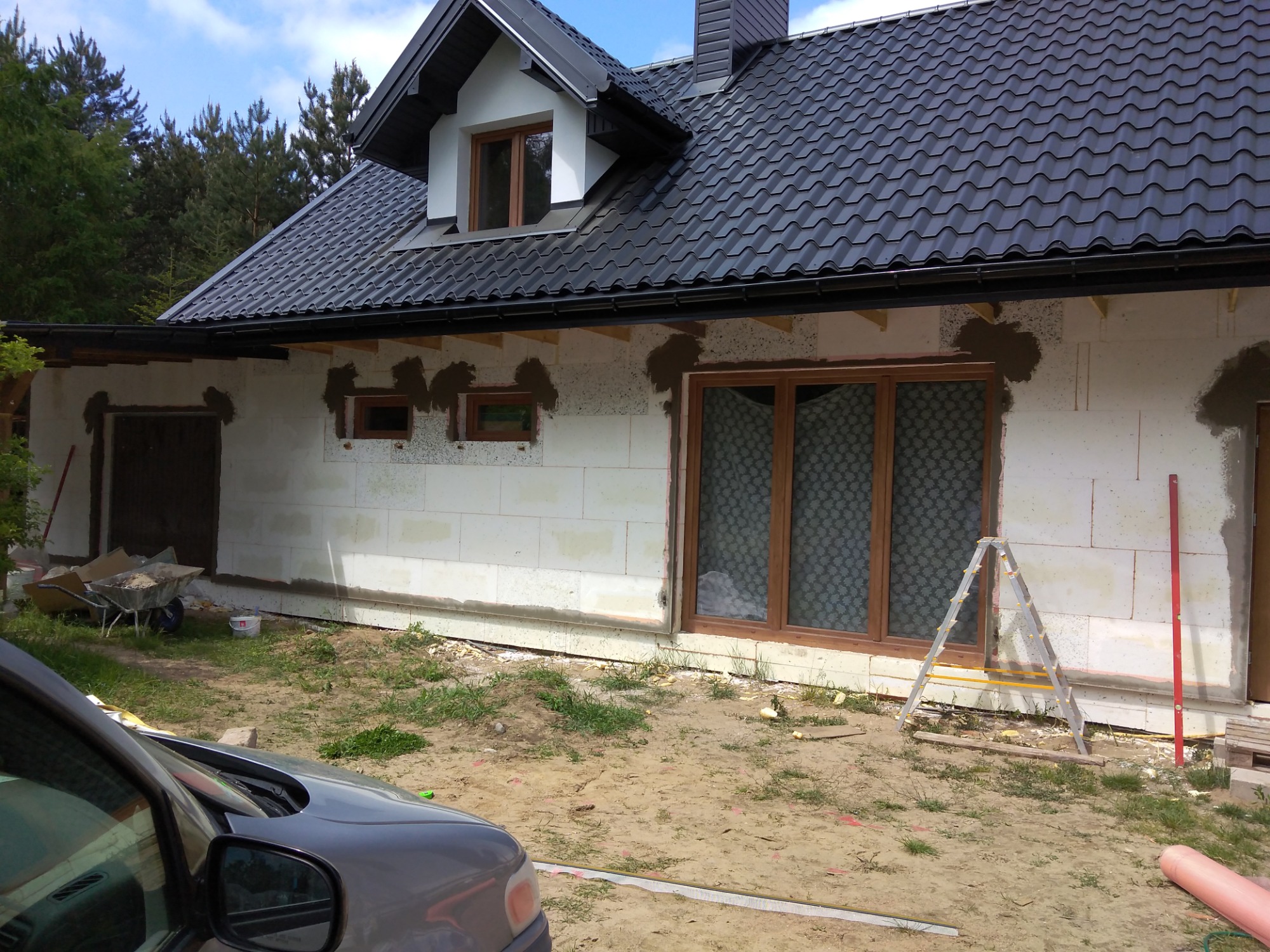 Dom w trakcie budowy: widoczne białe ściany z betonu komórkowego, zamontowane okna z drewnianymi ramami, czarny dach z dachówki ceramicznej, drabina oparta o ścianę, taczka z piaskiem w tle.
