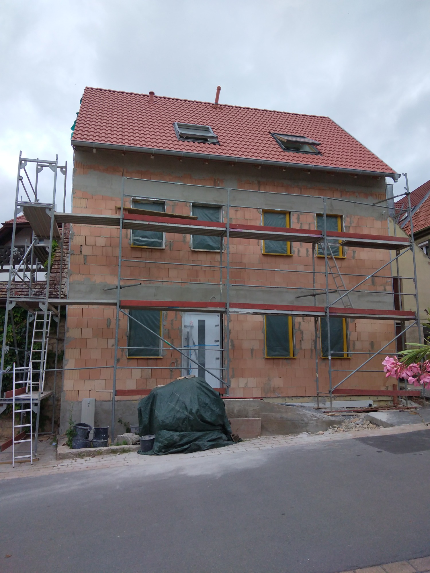 Dwupiętrowy dom z czerwonej cegły w trakcie budowy lub renowacji, z rusztowaniami, zabezpieczonymi oknami i nowym dachem z dachówki ceramicznej. Część elewacji pokryta szarym tynkiem.