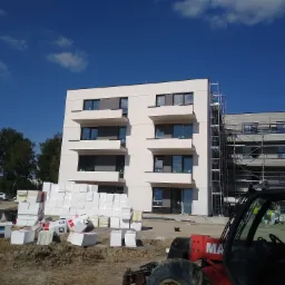 Nowoczesny budynek wielorodzinny w trakcie ocieplania, widoczne białe płyty styropianowe składowane przed budynkiem, w tle rusztowania i maszyna budowlana.