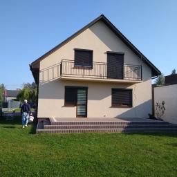 Tombud - Dwukondygnacyjny dom z beżową elewacją, brązowymi roletami i balkonem, widok z ogrodu z osobą niosącą wiadro z farbą