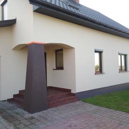 Tombud - Jasna elewacja domu jednorodzinnego z brązowymi akcentami: kolumna wejściowa i okna z roletami zewnętrznymi, widoczne schody wejściowe z płytek.