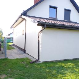 Tombud - Jasna elewacja domu jednorodzinnego z brązowymi rynnami i okiennicami, widok z boku na tle zielonego trawnika i sąsiednich budynków.