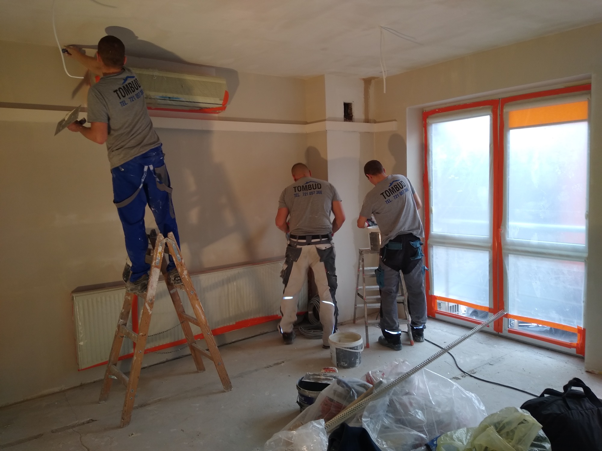 Trzech pracowników w szarych koszulkach firmy TOMBUD wyrównuje ściany wewnątrz pomieszczenia. Jeden stoi na drewnianej drabinie, drugi przy grzejniku, a trzeci przy oknie zabezpieczonym folią.