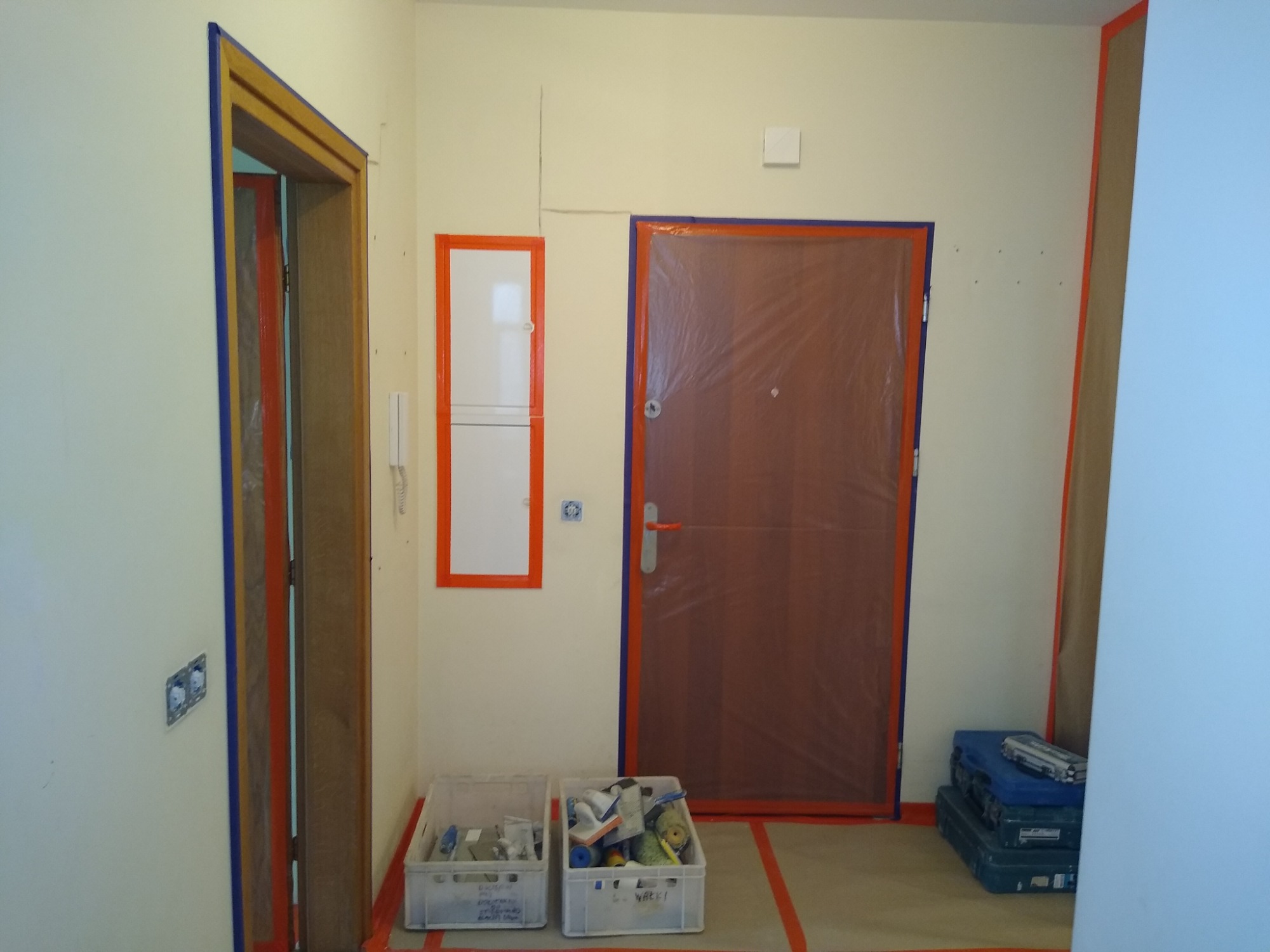 Przygotowanie do malowania wnętrza: zabezpieczone folią drzwi wejściowe, ościeżnice oklejone taśmą malarską, skrzynki z narzędziami i akcesoriami malarskimi.