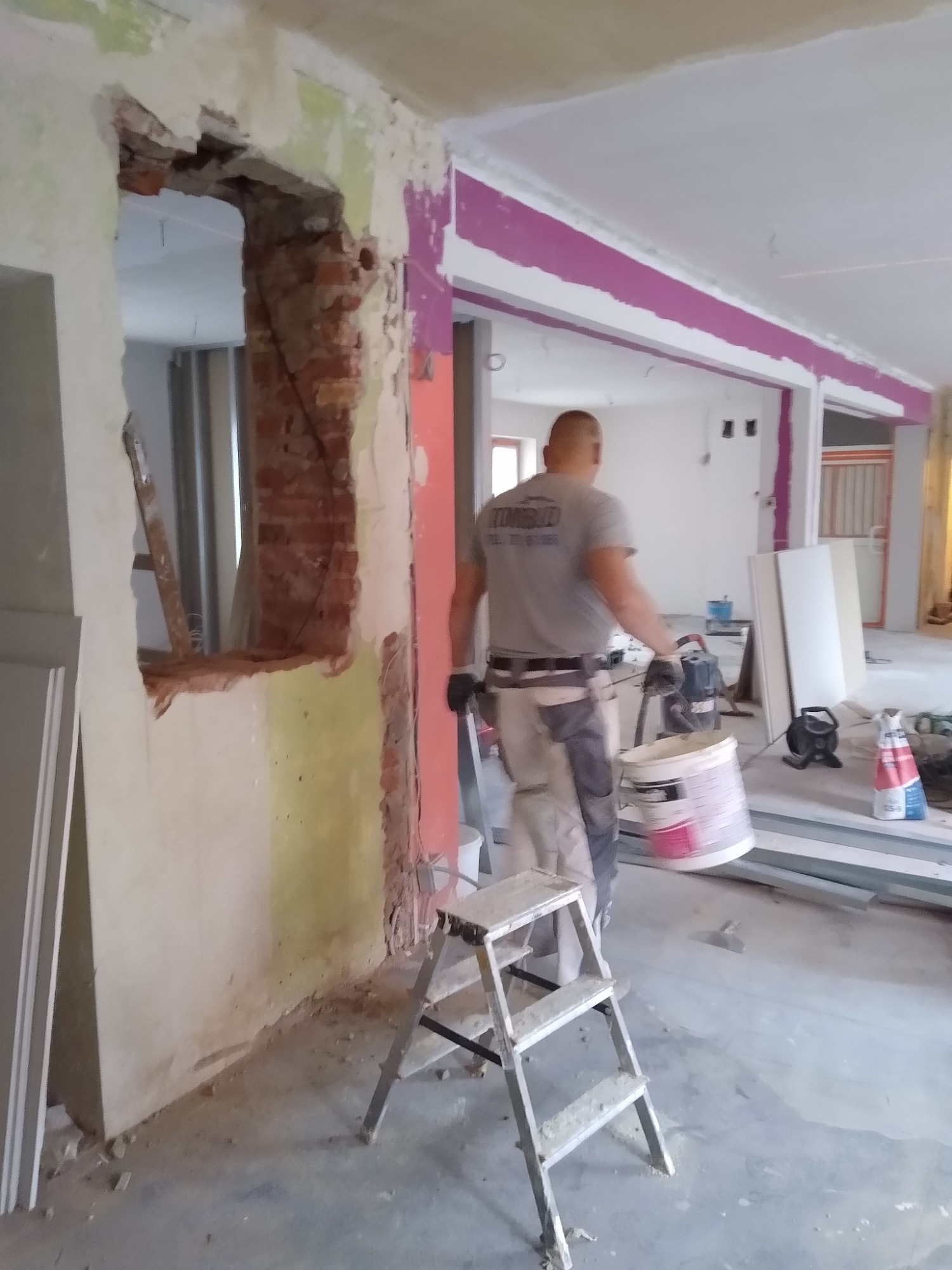 Wnętrze w trakcie remontu: murarz z wiadrem farby przechodzi obok ściany z odsłoniętą cegłą i resztkami starej farby, widoczny fragment schodków malarskich i listwy przypodłogowe.