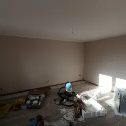 Pokój po malowaniu ścian na jasny beż, zabezpieczony folią malarską, z widocznym odkurzaczem przemysłowym, puszkami z farbą i elementami sufitu napinanego w folii.