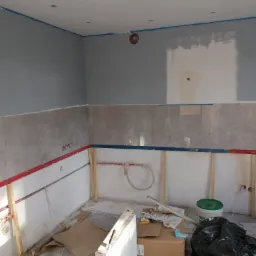 Wnętrze kuchni w trakcie prac wykończeniowych: częściowo położone płytki, ściany przygotowane do malowania z niebieską taśmą malarską, poziomice w kolorach czerwonym i niebieskim, widoczne...