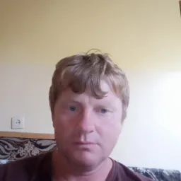 Portret mężczyzny z bliska, widoczny fragment sofy z wzorzystą tapicerką i gniazdko elektryczne na ścianie w tle.