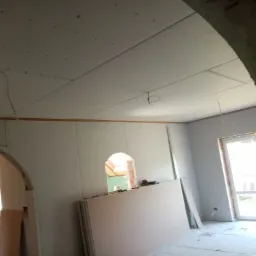 Wnętrze w trakcie remontu: zamontowane płyty kartonowo-gipsowe na suficie, widoczne kable, przygotowane płyty na podłodze, surowe wykończenie łukowego przejścia.