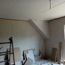 Wnętrze pokoju na poddaszu w trakcie prac wykończeniowych, widoczne płyty kartonowo-gipsowe na ścianach i suficie, drabina, narzędzia i materiały budowlane, okno z widokiem na zieleń.