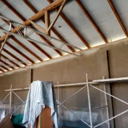 Surowe wnętrze budynku w trakcie prac wykończeniowych, widoczne świeżo nałożone warstwy tynku na ścianach, metalowe rusztowanie i drewniana konstrukcja dachu.