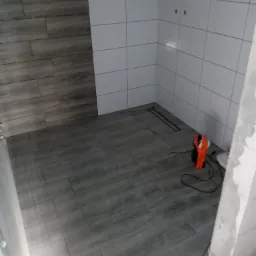 Wnętrze łazienki w trakcie remontu: ułożone płytki podłogowe imitujące drewno, białe płytki na ścianach, widoczny odpływ liniowy i nagrzewnica elektryczna.