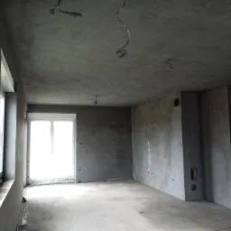 Surowe wnętrze w trakcie prac wykończeniowych, ściany i sufit pokryte świeżo nałożoną szarą warstwą, widoczne wystające przewody elektryczne i niedokończone otwory okienne.