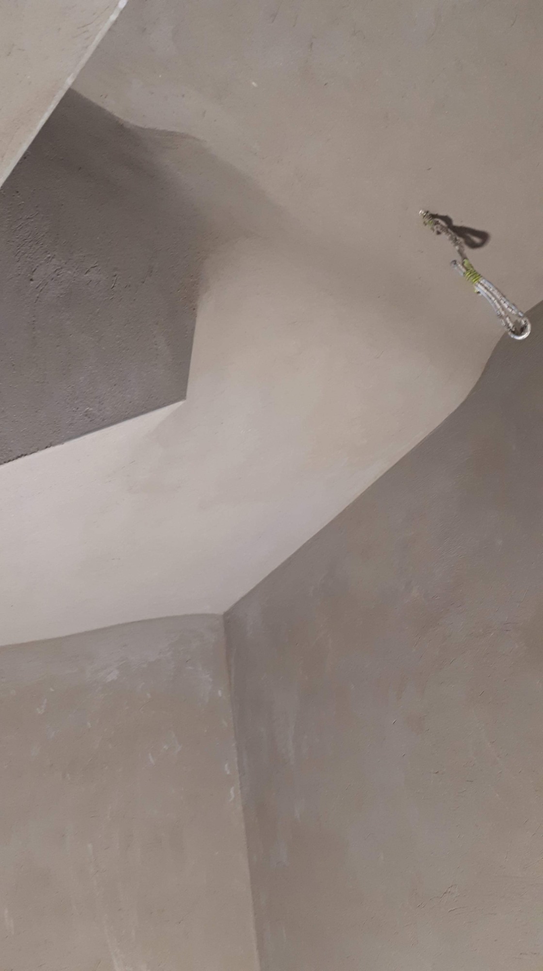 Szare, surowe ściany i sufit w trakcie tynkowania, widoczne nierówności i wiszący element mocujący.