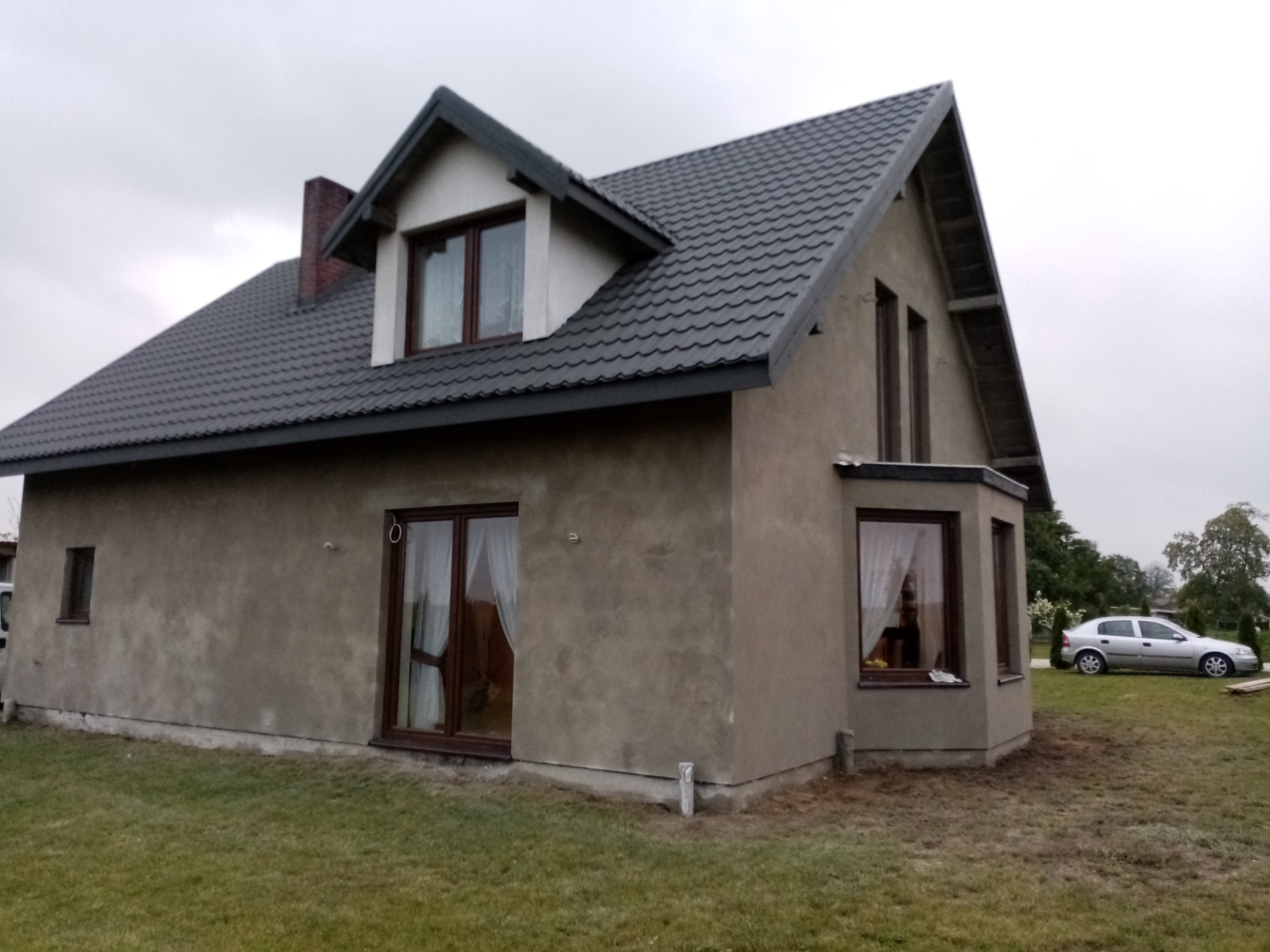 Dom jednorodzinny po tynkowaniu elewacji zewnętrznej, widoczna szara, surowa powierzchnia tynku przed malowaniem, z ciemną dachówką i brązowymi ramami okiennymi.