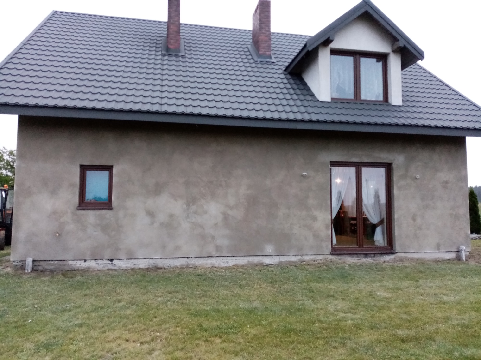Surowa elewacja domu jednorodzinnego z szarą dachówką, widoczne okna z brązowymi ramami i fragment traktora po lewej stronie budynku.