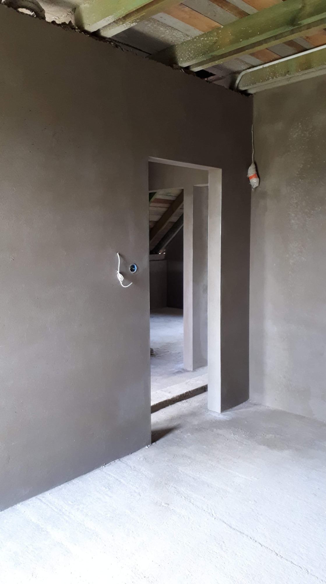 Wnętrze po tynkowaniu maszynowym z widocznymi przewodami elektrycznymi wystającymi ze ściany i surowym drewnianym stropem.